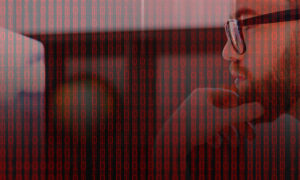 Profile view of man scratching chin while looking at a computer screen, superimposed with cyber security red binary numbers