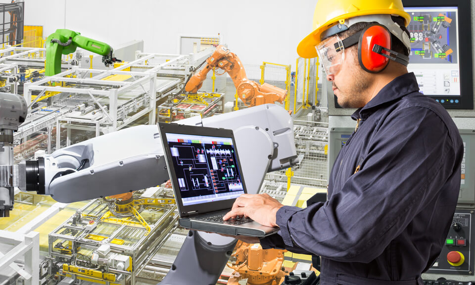 Male factory worker wearing a hard hat, working on a laptop beside an automated assembly-line