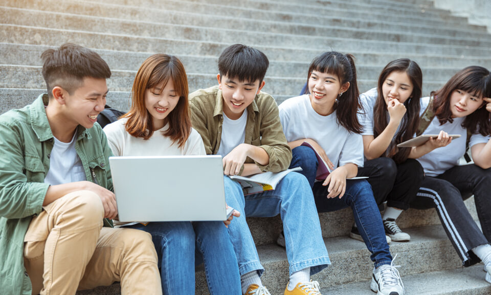 Asian students sitting in a row on concrete steps, using wireless devices